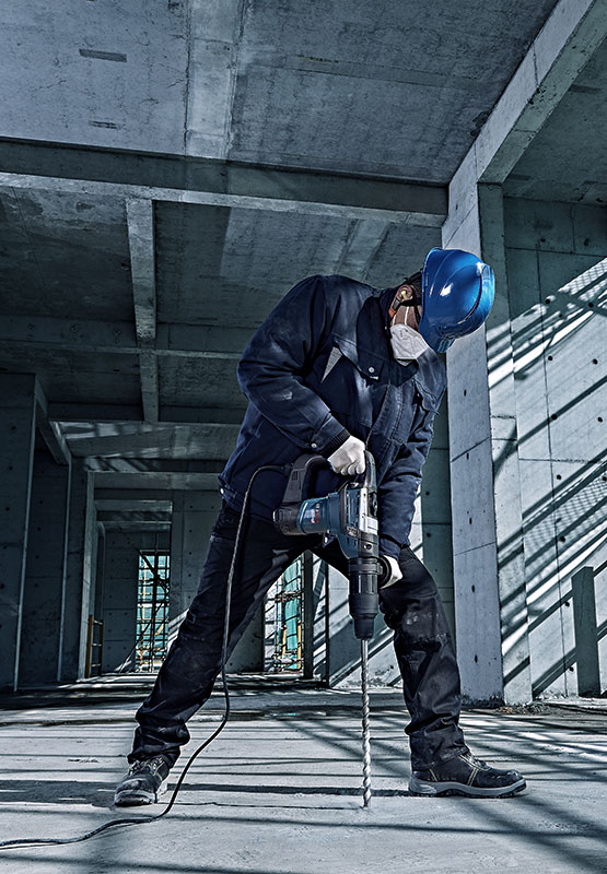 一名戴著藍色安全帽和口罩的工人在混凝土房間裡鑽孔。